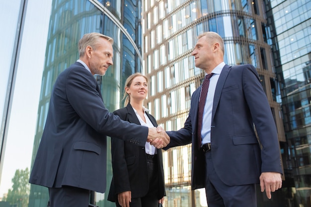 Free photo confident middle-aged businessmen greeting each other and handshaking. successful content young businesswoman standing near them and smiling. building on background. teamwork and partnership concept