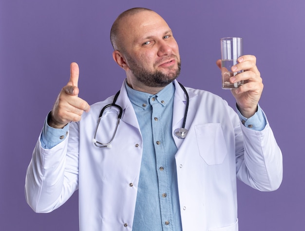 Confident middle-aged male doctor wearing medical robe and stethoscope holding glass of water