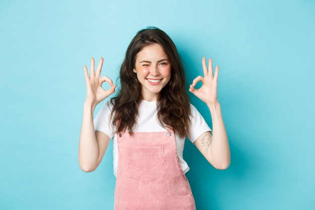 Confident and positive young woman winking and smiling, showing okay signs in approval, say yes, give approval, praise good work, standing against blue background.