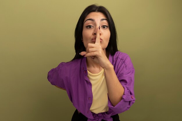 Confident pretty brunette woman doing silence gesture isolated on olive green wall