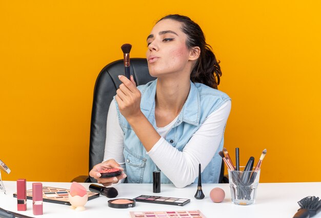 Confident pretty caucasian woman sitting at table with makeup tools holding blush and looking at makeup brush 