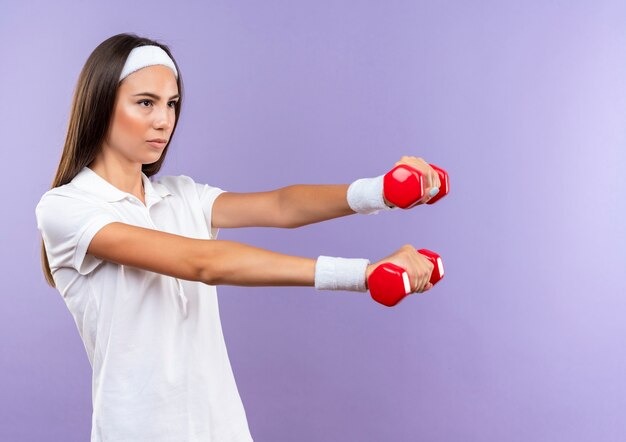 Confident pretty sporty girl wearing headband and wristband holding dumbbells isolated on purple wall with copy space