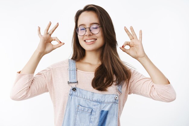 Confident and satisfied young female customer showing raised okay gesture and winking in approval and like smiling broadly pleased with good quality of product on gray.