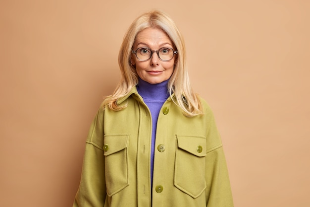 Free photo confident senior female teacher dresses for work has blonde hair wears transparent glasses turtleneck and stylish jacket going to have outdoor walk during autumn day.