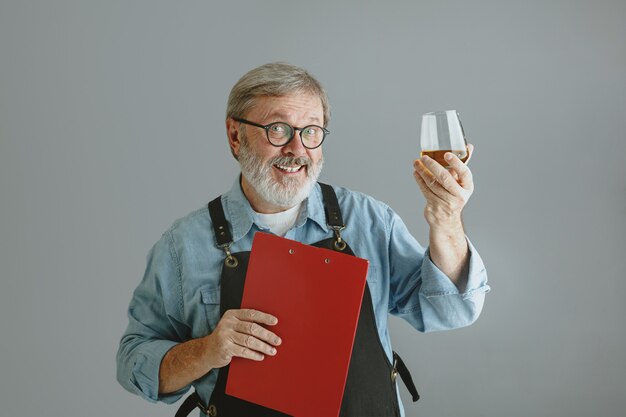 Confident senior man brewer with self crafted beer in glass on wooden barrel on grey background.