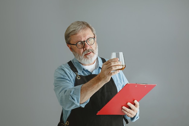 Confident senior man brewer with self crafted beer in glass on wooden barrel on grey wall