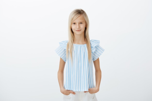 Free photo confident serious-looking blond girl in blue clothes, holding hands in pockets and smiling broadly while on children audition, being self-assured and relaxed over gray wall