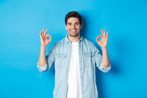 Free photo confident smiling adult man, showing ok signs and looking pleased, like something, standing against blue background.