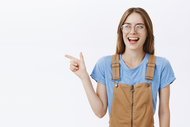 Free photo confident smiling pretty girl student in glasses looking happy, pointing finger left at your logo