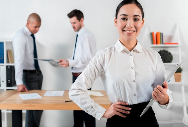 Confident smiling young businesswoman holding clipboard in hand with two businessman standing at backdrop