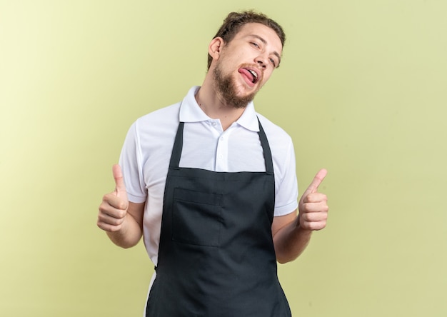 Free photo confident tilting head young male barber wearing uniform showing thumbs up isolated on olive green wall