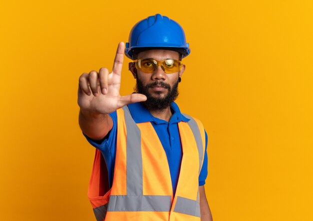 Confident young afro-american builder man in safety glasses wearing uniform with safety helmet pointing up isolated on orange wall with copy space