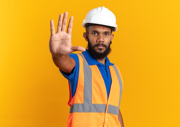 Confident young afro-american builder man in uniform with safety helmet doing stop gesture isolated on orange background with copy space