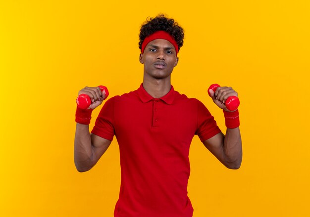 Confident young afro-american sporty man wearing headband and wristband exercising with dumbbell isolated on yellow wall