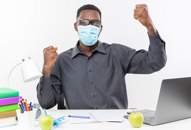 Confident young afro-american student in optical glasses wearing medical mask sitting at desk with school tools raising fists up