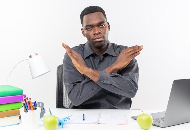 Confident young afro-american student sitting at desk with school tools crossing his hands gesturing no sign 