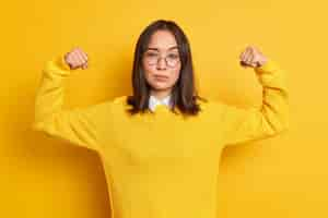 Free photo confident young asian woman shows arm muscles feels like hero demonstrates her power and strength looks seriously wears round optical glasses sweater.