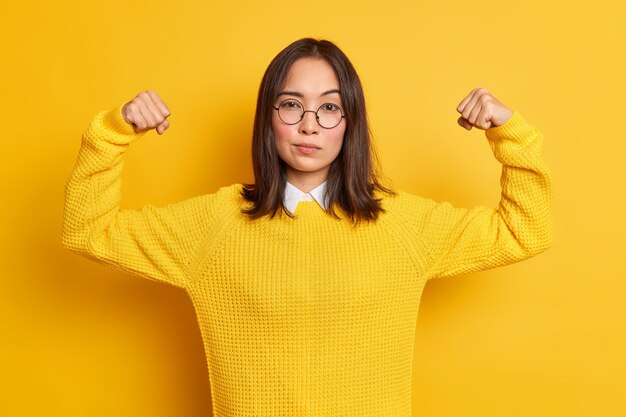 Confident young Asian woman shows arm muscles feels like hero demonstrates her power and strength looks seriously wears round optical glasses sweater.