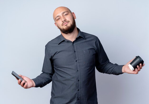 Confident young bald call center man holding mobile phone and plastic coffee cup isolated on white 