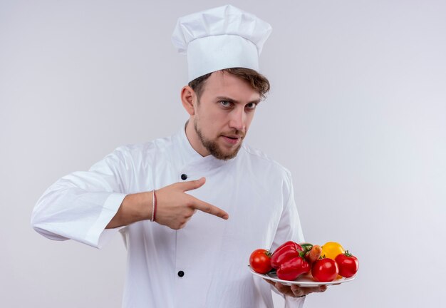 A confident young bearded chef man wearing white cooker uniform and hat pointing at a white plate with fresh vegetables on a white wall