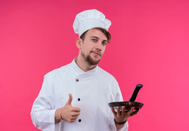 A confident young bearded chef man in white uniform showing thumbs up with frying pan in the other hand while looking on a pink wall