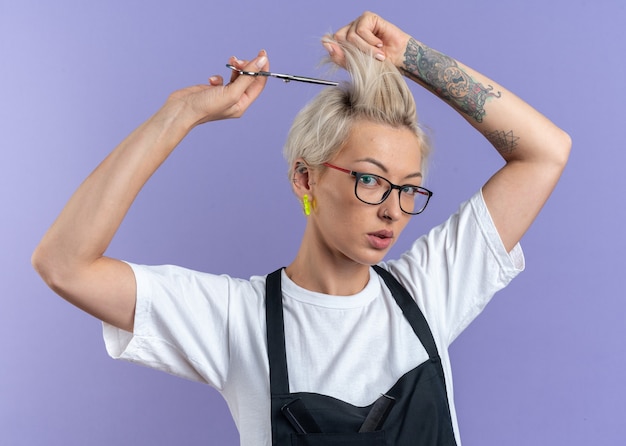 Free photo confident young beautiful female barber in uniform wearing glasses cutting hair with scissors isolated on blue wall