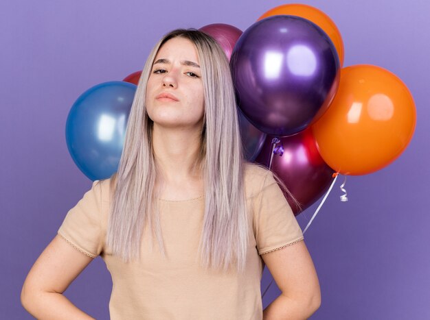 Confident young beautiful girl standing in front balloons isolated on blue wall