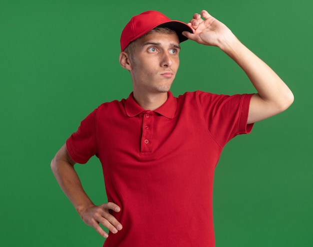 Free photo confident young blonde delivery boy puts hand on cap and looks at side isolated on green wall with copy space