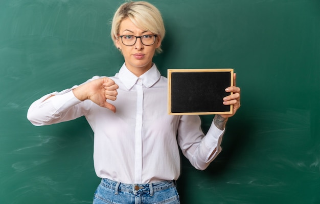 confident young blonde female teacher wearing glasses in classroom standing in front of chalkboard showing mini blackboard looking at front showing thumb down