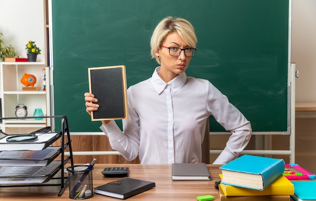 Confident young blonde female teacher wearing glasses sitting at desk with school tools in classroom showing mini blackboard keeping hand on waist looking at camera