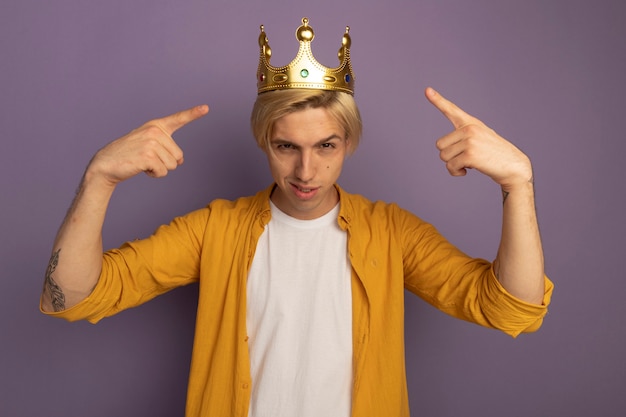 Confident young blonde guy wearing yellow t-shirt and crown points at himself isolated on purple