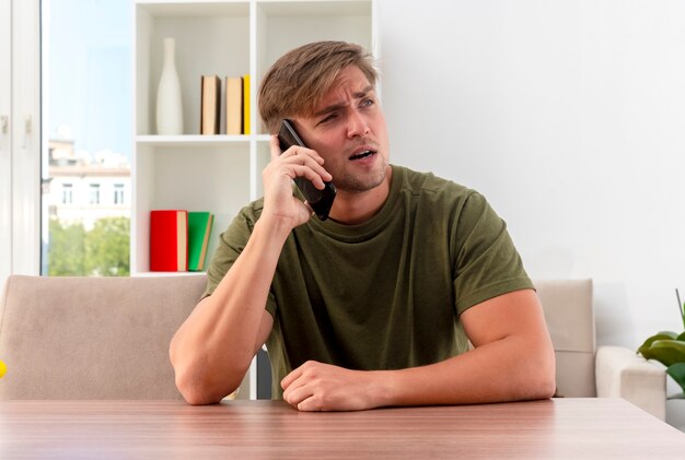 Confident young blonde handsome man sits at table talking on phone and looking at side