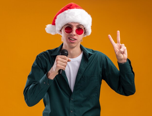 Free Photo confident young blonde man wearing santa hat and glasses talking into microphone looking at camera doing peace sign isolated on orange background