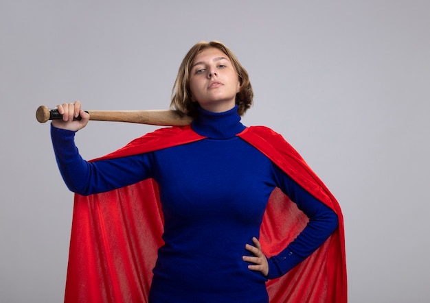 Free photo confident young blonde superwoman in red cape holding baseball bat keeping hand on waist looking at front isolated on white wall