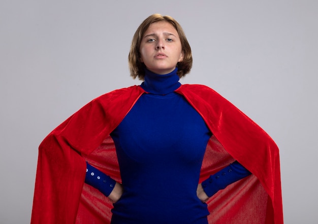 Free photo confident young blonde superwoman in red cape keeping hands on waist looking at front isolated on white wall