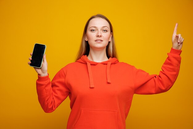 Confident young blonde woman holding mobile phone pointing up 