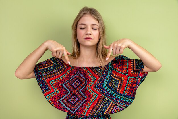 Confident young blonde woman pointing down standing with closed eyes 