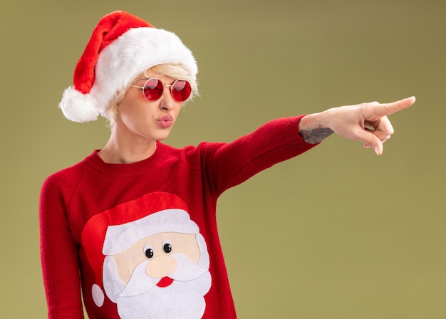 Free photo confident young blonde woman wearing christmas hat and santa claus christmas sweater with glasses looking and pointing at side with pursed lips isolated on olive green background