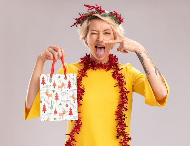 confident young blonde woman wearing christmas head wreath and tinsel garland around neck holding christmas gift bag looking  showing v-sign symbol near eye winking showing tongue isolated on white wall