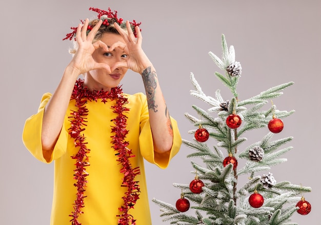 Free photo confident young blonde woman wearing christmas head wreath and tinsel garland around neck standing near decorated christmas tree looking  doing heart sign in front of face isolated on white wall