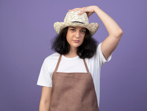 Confident young brunette female gardener in uniform wearing and putting hand on gardening hat isolated on purple wall