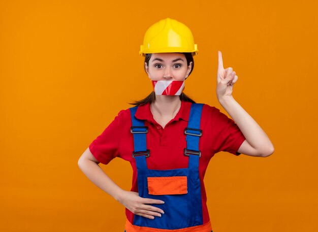 Confident young builder girl mouth sealed with warning tape and points up on isolated orange background