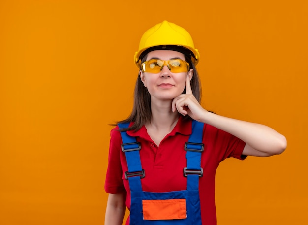 Free photo confident young builder girl with safety glasses puts hand on chin and looks up on isolated orange background with copy space