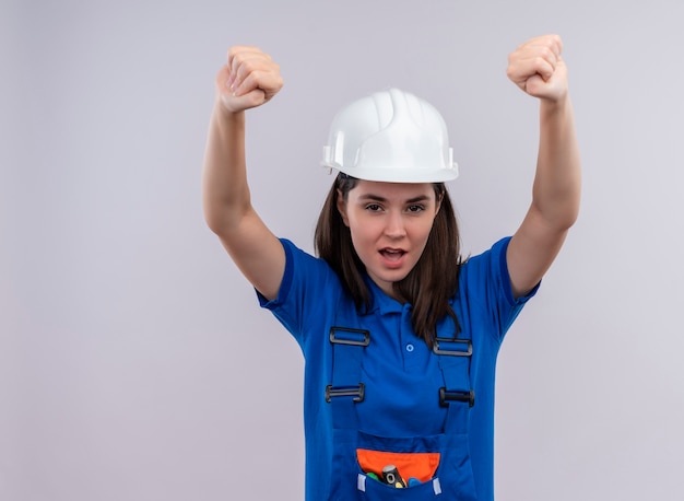 Confident young builder girl with white safety helmet and blue uniform raises fists up and looks at camera on isolated white background with copy space