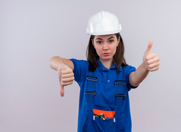 Confident young builder girl with white safety helmet and blue uniform thumbs down and thumbs up on isolated white background with copy space