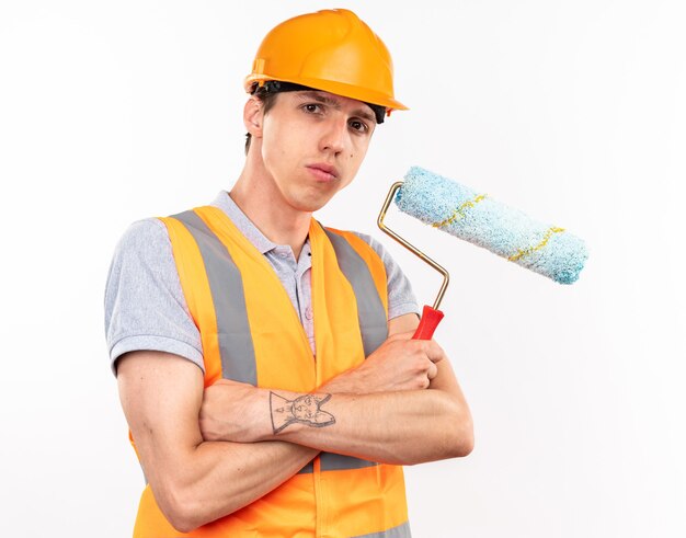 Confident young builder man in uniform crossing hands holding roller brush 