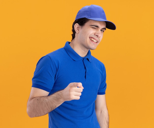 Confident young caucasian delivery man in blue uniform and cap winking looking and pointing at camera solated on orange background