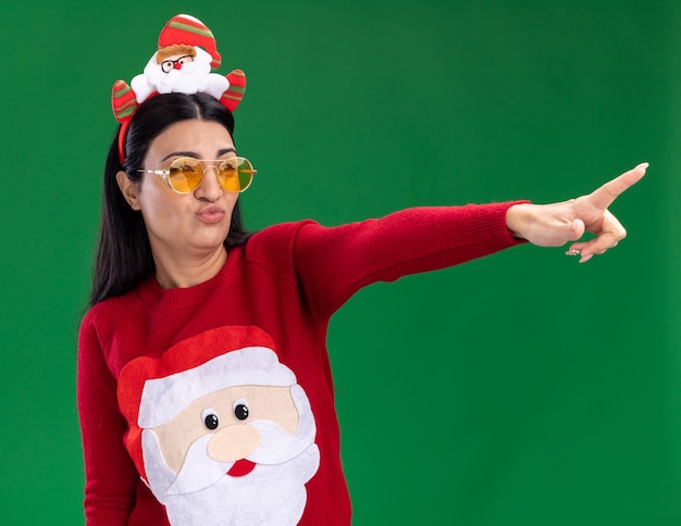 Free photo confident young caucasian girl wearing santa claus headband and sweater with glasses looking and pointing at side with pursed lips isolated on green background