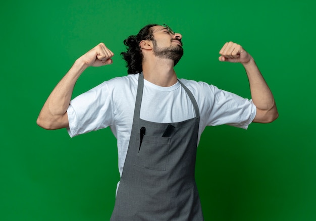 Confident young caucasian male barber wearing glasses and wavy hair band in uniform gesturing strong with closed eyes isolated on green background with copy space