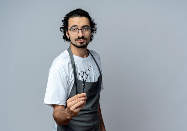 Confident young caucasian male barber wearing glasses and wavy hair band in uniform stretching out comb and scissors at camera isolated on white background with copy space